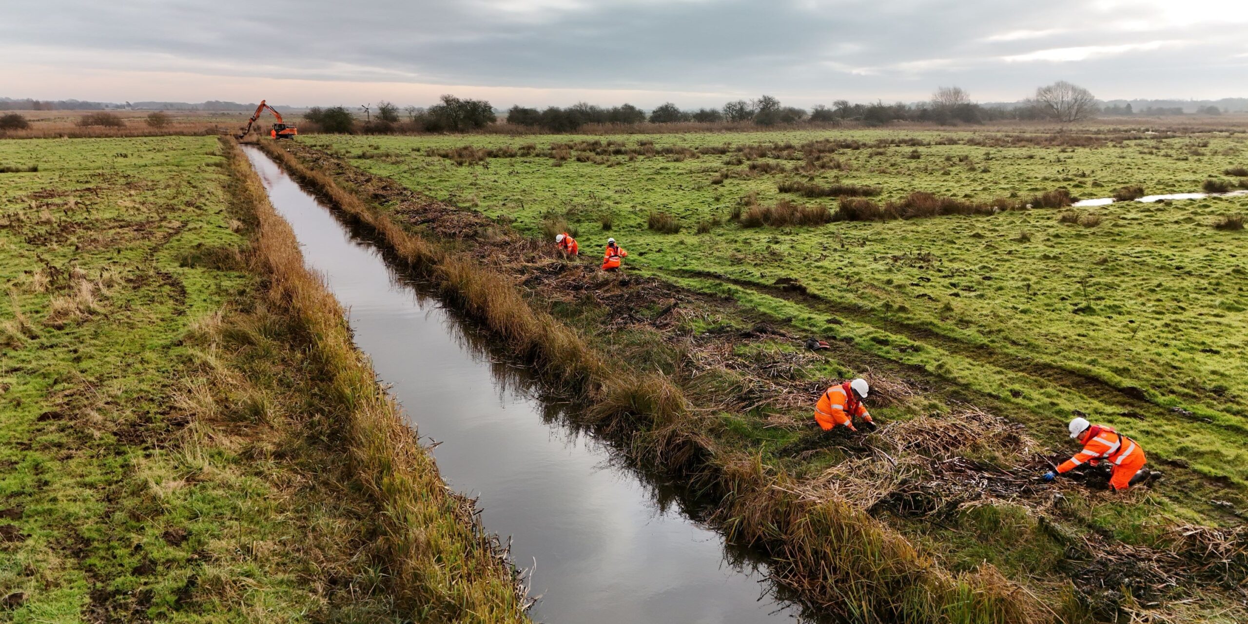 Panoramic Excavator carrying out watercourse maintenance as part of the Grasswrack Pondweed rescue - environment team seen in foregound collecting plants October 2024