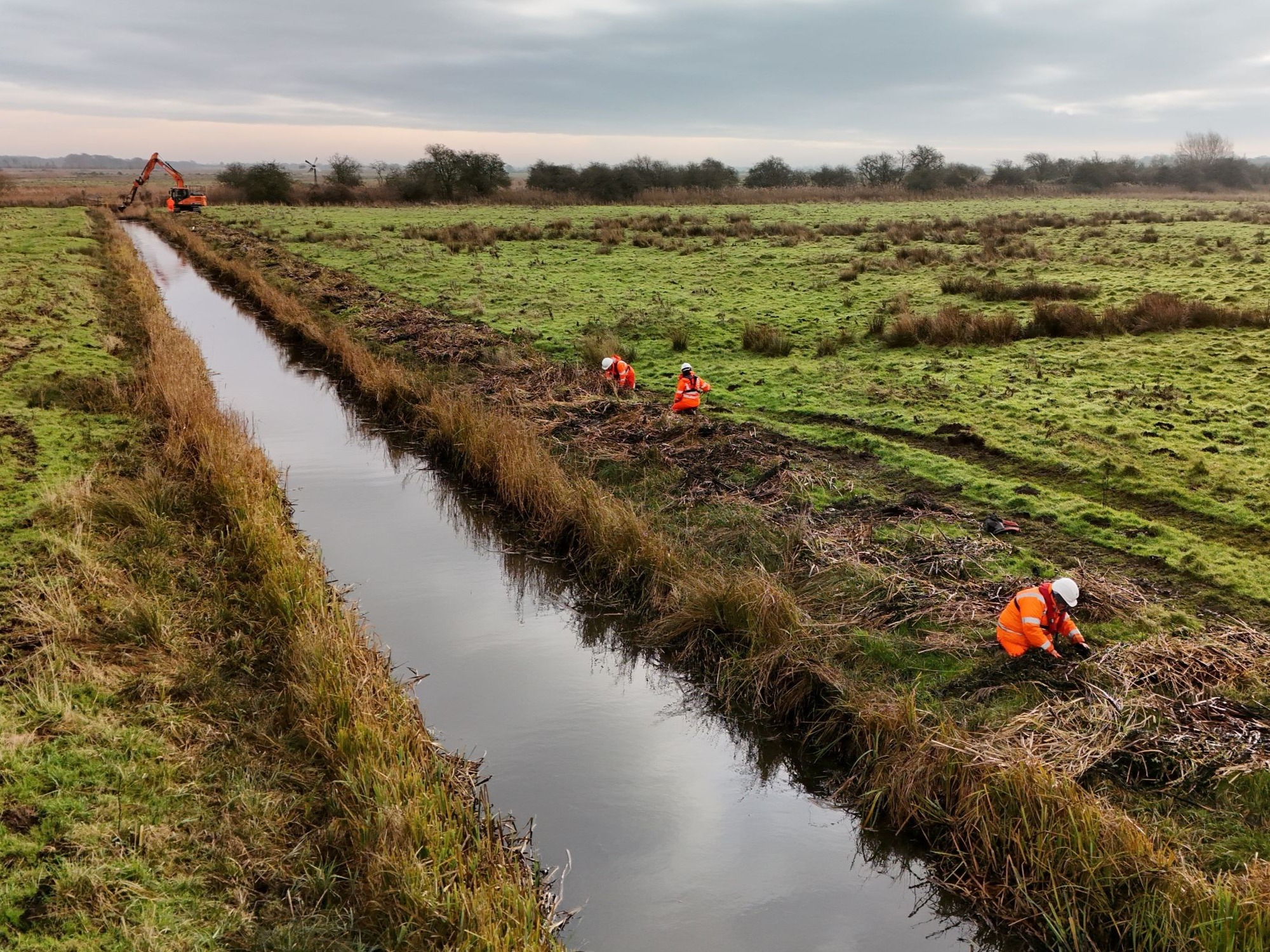 Excavator carrying out watercourse maintenance as part of the Grasswrack Pondweed rescue - environment team seen in foregound collecting plants October 2024